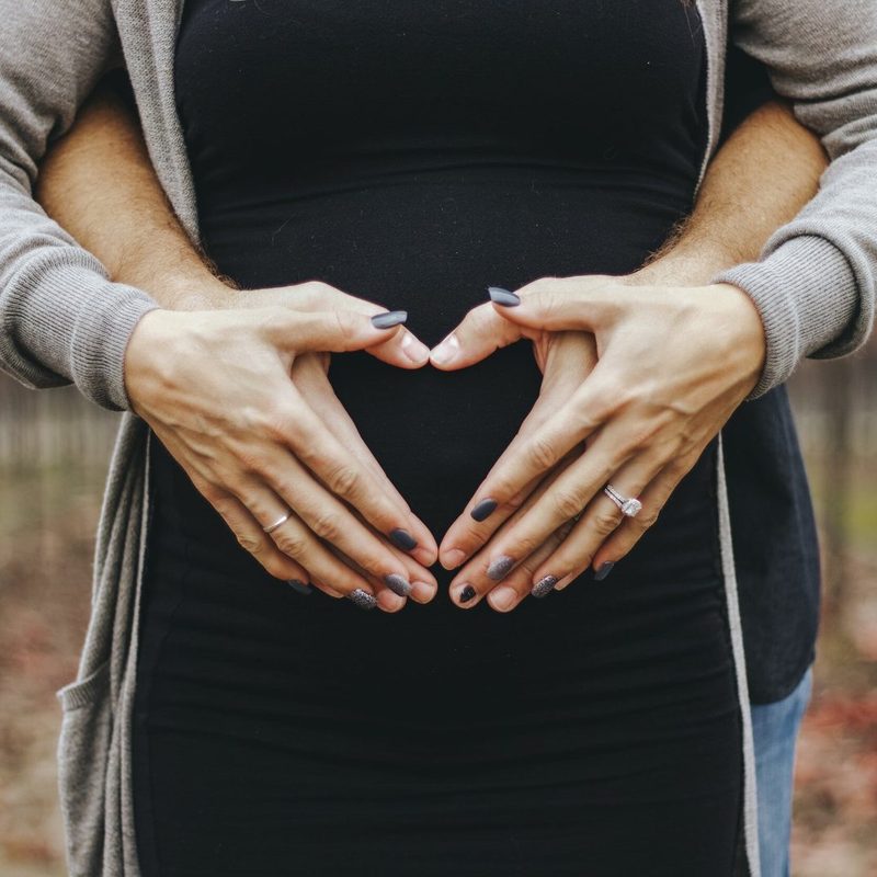 Pregnant woman making a heart shape with her hands over her bump while her partner holds her