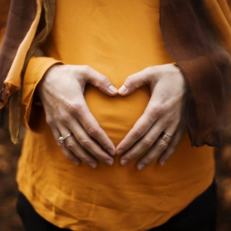 Hands forming a heart over a pregnant bump in a mustard jumper
